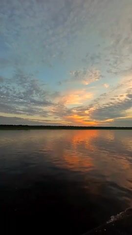The video shows a wide shot of a calm body of water, likely a river or lake, at sunset. The sky is filled with scattered clouds that are illuminated by the setting sun, creating a gradient of colors from pale blue to orange and pink. The sun itself is a bright orange glow on the horizon, casting a reflection on the water's surface. A dark, distant treeline or shoreline is visible across the water. In the foreground, the edge of a dark object, possibly a boat or dock, is visible at the bottom right of the frame. The water is dark and reflects the colors of the sky, creating a shimmering effect. The scene is peaceful and serene.