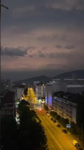 A vertical video shows a city street at dusk or night. The sky is dark and cloudy, with some hints of pink and purple from the setting sun. Streetlights and building lights illuminate the scene, casting a warm glow on the wet asphalt road. Cars with their headlights and taillights on are driving down the street. Buildings of various heights and architectural styles line both sides of the road. In the distance, mountains are visible under the moody sky. The overall atmosphere is calm and slightly melancholic. There are no people clearly visible.