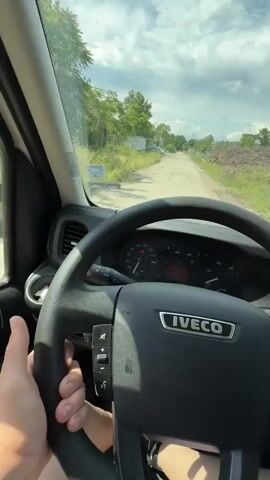 The video provides a first-person view from inside an IVECO vehicle, likely a van or truck, as it is driven along an unpaved road in Kalitinovo, Bulgaria. The driver's hands are visible at the bottom left, holding the steering wheel which prominently displays the "IVECO" logo and various control buttons. The dashboard is visible behind the steering wheel, with the digital display showing "15:31". Through the windshield, a rough, dusty dirt road stretches ahead, flanked by green trees and bushes on both sides. On the left, a fence and some distant structures are partially visible, while on the right, piles of debris or rubble line the roadside. The sky is partly cloudy, indicating an early afternoon setting. A phone mount is attached to the right side of the dashboard.