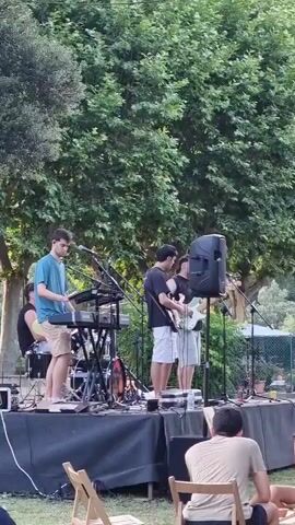 A band performs on an outdoor stage. The band consists of four young men. One plays a keyboard, another a guitar, and a third a bass guitar. A fourth person is partially obscured and appears to be playing drums. They are set up on a black stage covered with what appears to be a tarp. There are microphones and speakers on the stage. In the foreground, several people are seated on chairs watching the performance. They are on a grassy area. Behind the stage, there are trees with lush green foliage, and a fence can be seen in the distance. The lighting suggests it is daytime, possibly late afternoon or early evening.