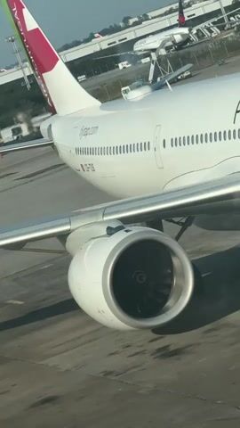 A vertical video shows an airplane's fuselage, wing, and engine from a high angle. The white airplane has "PORTUGAL" written on its side in black letters. The engine is visible with its fan blades stationary. In the background, other airplanes and airport structures are faintly visible under a sunny sky.