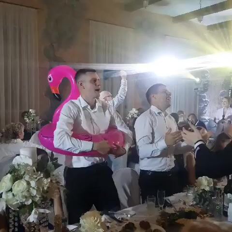The video shows a wedding reception. Two men are in the foreground, one is wearing a pink flamingo inflatable around his torso. Both men are wearing white shirts and black pants, and they appear to be singing or speaking to the crowd. They are standing at a table laden with food and drinks, with other guests visible in the background. A bright spotlight shines down on the men, illuminating the scene. The overall atmosphere appears to be festive and celebratory.