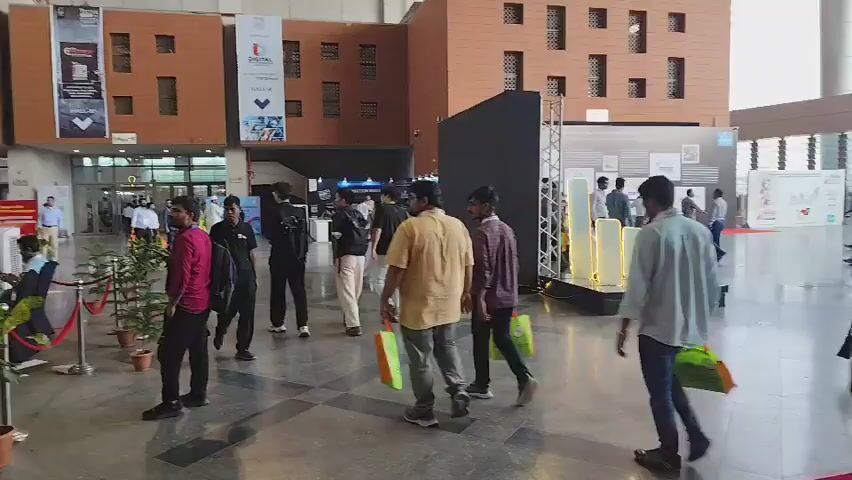 The video begins by showing a bustling indoor space, possibly a convention center or exhibition hall, with people walking around. A red carpet is laid out on the floor, indicating a special event. Several individuals are seen carrying green shopping bags, suggesting they are attending a fair or expo. The architecture of the building features large windows and prominent brickwork. In the background, there are booths and displays set up, hinting at various exhibitions or presentations. The camera pans to show more of the crowd and the layout of the venue, with some people engaged in conversations and others browsing the exhibits. Towards the end, the video focuses on some individuals seated at a desk with computers, possibly working at an information counter or registration area. The overall atmosphere appears to be one of a lively and organized event.