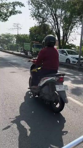 The media file depicts a person riding a scooter on a paved road. The rider is wearing a maroon long-sleeved shirt, black pants, and a black helmet. They are positioned in the center of the frame, moving away from the viewer. The scooter is white and black with yellow accents, and has a visible license plate with the text "W 2957 NAP 08-26".

The scene is outdoors, likely during the daytime, as indicated by the bright sunlight casting long shadows. The weather appears to be clear and sunny. The road is bordered by a sidewalk or embankment on the right, with trees and vegetation. In the background, several vehicles are visible, including a green truck and a white car. A power pole is also present on the right side of the frame. The environment suggests an urban or semi-urban setting, consistent with the provided location context of Siring, East Java. There are no explicit emotional cues visible from the rider. The overall impression is a mundane street scene on a bright day.