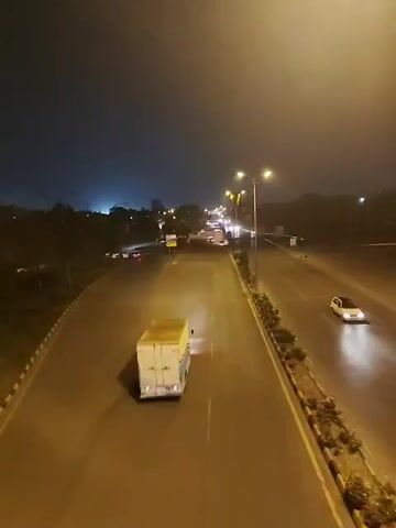 The video depicts a multi-lane road at night, illuminated by streetlights. The road appears wet, possibly from rain. Several vehicles, including a white box truck and cars, are moving along the road. There is a motorcycle. The surrounding environment includes trees and greenery along the roadside. In the distance, there are city lights. The sky is dark. The video appears to be taken from an elevated position, possibly a bridge or overpass.
