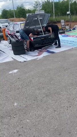 The video shows two men working on the engine of a car with its hood open. They are both wearing casual clothing. One man is wearing a black t-shirt and blue denim shorts, and the other is wearing a black t-shirt. They are bent over the engine, appearing to inspect or adjust something. The car is parked on a large white tarp, which has some graphics on it. In the background, there are other vehicles, trees, and some piles of gravel. The ground appears to be asphalt. A third person is partially visible in the foreground, facing away from the car.