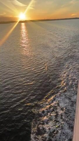 A wide shot of a body of water at sunset. The sun is low on the horizon, casting long rays across the water. There are some gentle waves and a wake from a boat or ship on the right side of the frame. The sky is a mix of orange, yellow, and blue. The overall mood is peaceful and serene.