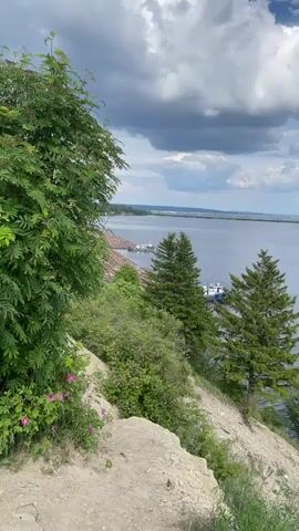 The video presents a panoramic view from an elevated, rocky and dirt-covered cliff overlooking a large body of water, likely a river or lake, under a cloudy sky. The camera pans slowly from left to right, capturing the expansive natural landscape.

In the foreground on the left, lush green trees and bushes, including one with pink flowers, are visible. The slope down to the water is steep and covered with sparse vegetation and exposed earth. Further down the slope and at the water's edge, more trees, including two prominent evergreen trees, punctuate the scene.

On the water, several small white and blue boats are docked near the shore, and the water surface shows ripples, reflecting the light. In the distance, a faint outline of a bridge or landmass stretches across the horizon. The sky is partly cloudy, with both fluffy white clouds and darker grey clouds suggesting dynamic weather.

A child's voice is audible in the background, adding a lively, though unseen, element to the peaceful natural vista. The scene appears to be set during the daytime, offering a wide, scenic view of the water and distant shore. There is no visible text in the video.