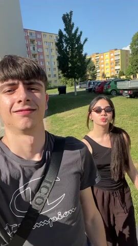 This is a selfie-style video clip taken outdoors on a sunny day. The foreground features a young man with dark, bowl-cut hair and a grey t-shirt with a white Champion logo. He is smiling and looking directly at the camera, with a black strap of a bag crossing his chest. Behind him, a young woman with long, dark hair and sunglasses is partially visible. She is wearing a black tank top and dark pants and is holding a white plastic bag.

The background depicts a residential area with a multi-story apartment building, characterized by colorful facades and rows of windows. A patch of green grass and trees are also present, along with parked cars and a small truck. The clear blue sky and bright sunlight suggest it is daytime. The overall mood appears light and cheerful, with both individuals smiling.

There is visible text on the man's t-shirt: "Champion". The timestamp on the video indicates it was recorded on July 24, 2023. The location context provided is Dobřany, Czechia.