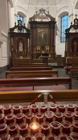 The video shows the interior of a church. In the foreground, there are rows of red votive candles in glass holders, with one candle lit. Behind the candles are rows of wooden pews with red seats and backs. Further back, at the front of the church, is an altar area. There is a large wooden altarpiece with a painting of the crucifixion of Jesus in the center. There are also statues and other religious artifacts visible. The church has high ceilings and large windows. The lighting is dim, creating a somber atmosphere. There are no people visible in the video.