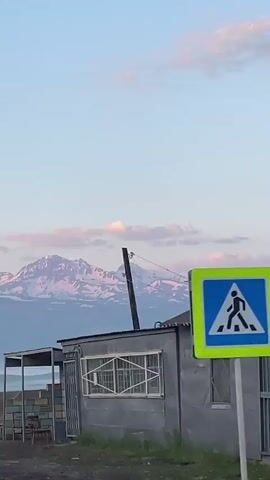 The video shows a landscape scene with mountains in the background. In the foreground, there are some buildings and structures, including a roofed shelter with chairs under it, and a grey building with a window and a metal gate. A utility pole with wires is also visible. The sky is a pale blue with some pinkish clouds, and the mountain peaks are capped with snow, also tinged with pink from the light. A misty valley can be seen between the foreground buildings and the mountains. The overall impression is a rural or semi-rural setting at dawn or dusk. There are no people visible in the video.