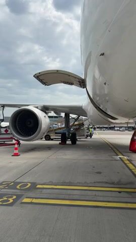 The video shows a close-up view of an airplane at an airport. The camera is positioned beneath the wing of the plane, capturing the landing gear, engine, and part of the fuselage. A cargo loader is parked near the plane, with a person wearing a light blue jumpsuit walking up the loader's stairs. The plane's engine is visible with its fan blades in detail. The landing gear has two sets of wheels, and a red chock is placed behind one of the wheels. The fuselage has an open cargo door. Yellow lines are painted on the airport tarmac. The sky is overcast, suggesting a cloudy day.