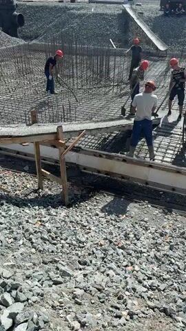 The video shows a construction site where workers are pouring concrete. Multiple individuals wearing hard hats are actively engaged in the process. They are using shovels to spread and level the wet concrete over a reinforced metal grid. A concrete chute is visible, indicating the source of the concrete being poured from a truck in the background. The environment appears to be an outdoor construction area with loose gravel and rebar present. The overall scene depicts a group of construction workers performing the essential task of concrete placement.
