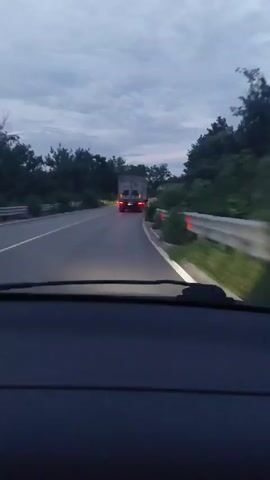 The video shows a view from inside a car driving on a road at dusk. The road is asphalt and appears to be wet. Trees line both sides of the road. In front of the car, a truck with its brake lights illuminated is visible on the road ahead. The sky is cloudy and the light is dim. The camera is pointed forward, showing the dashboard and windshield of the car.