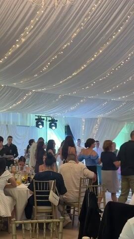 A vertical video captures people gathered in what appears to be a reception hall or event space. The room is decorated with white draped fabric on the ceiling, adorned with strings of small lights, and features a chandelier in the upper left. A group of people is standing and dancing in a line on the right side of the frame, facing away from the camera. Several individuals are seated at circular tables in the foreground, dining and conversing. A man in a light-colored shirt is seated facing away from the camera in the center foreground, while a woman with dark hair in a bun is seated to his left, looking toward the dancing group. In the background, stage lights are visible, indicating a performance area. The overall atmosphere suggests a lively social event.
