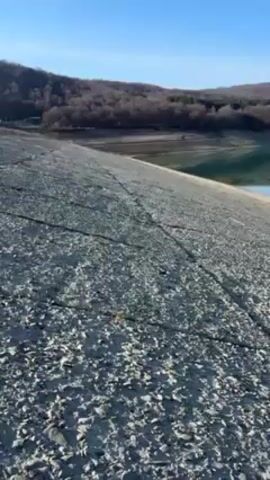 The video shows a tranquil landscape with a dam and a lake. The dam is made of gray, rough material, possibly concrete or stone, with visible cracks and lines running across its surface. The lake is calm and blue, reflecting the clear sky and the wooded hills on the opposite shore. The hills are covered with leafless trees, indicating it might be late autumn or early spring. The overall atmosphere is peaceful and serene, with the clear sky suggesting a bright, sunny day. The camera slowly pans across the scene, capturing the details of the dam, the lake, and the surrounding nature.