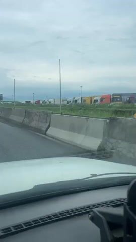 This video shows a view from the interior of a vehicle driving on a highway in Nogir, Russia. The foreground is dominated by the vehicle's dashboard and windshield. Beyond the windshield, a concrete barrier separates the highway lanes from a grassy area with parked semi-trailer trucks. The trucks are diverse in color, including white, red, yellow, and orange. The sky is overcast with clouds, suggesting a possibly cool or rainy day.

In the distance, there are more trucks and what appears to be a large digital billboard or sign. The highway itself has at least two lanes of traffic in the direction the camera is facing, and there are other vehicles visible, including cars and a dark-colored sedan in the left lane. Lampposts line the side of the highway, and distant hills or mountains are visible under the cloudy sky. The overall impression is a mundane highway scene, likely during the daytime, with a focus on commercial vehicles and infrastructure.