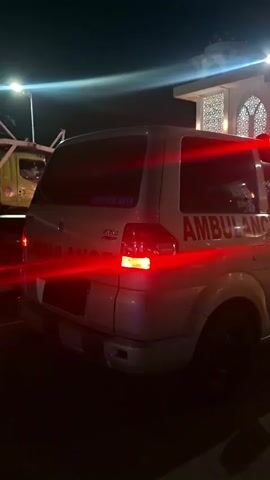 This is a video captured at night in Paya Tringgadeng, Indonesia. The primary subject is a white ambulance with red flashing lights, parked on a road. The ambulance has the word "AMBULANCE" written in red on its side. In the background, there's a building with architectural elements that suggest it might be a religious structure, possibly a mosque, with arched windows and decorative patterns. The lighting is dim, typical of nighttime, with strong artificial lights from street lamps and the ambulance itself casting shadows.

A tow truck is partially visible in the distance on the left side of the frame, indicating a potential incident or recovery operation. The scene appears to be somewhat chaotic due to the flashing lights and the presence of emergency or recovery vehicles. The weather seems clear, as there are no visible signs of rain or fog. The overall mood is one of urgency or possibly a response to an emergency.

Visible text on the ambulance includes "AMBULANCE" and a smaller blue sticker with text that is difficult to read clearly. On a yellow barrier in the background, text is visible, including "SE AT-TAQARRO". The time of day is clearly night.