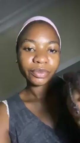 A young Black woman with a pink headband is holding a child in her arms while recording a video. The woman is wearing a gray tank top and has clear skin. The child's face is partially obscured by the woman's chest, but the child has dark hair in braids. The background is a dimly lit room with a ceiling visible at the top of the frame. Music is playing in the background. The woman is looking at the camera and talking.