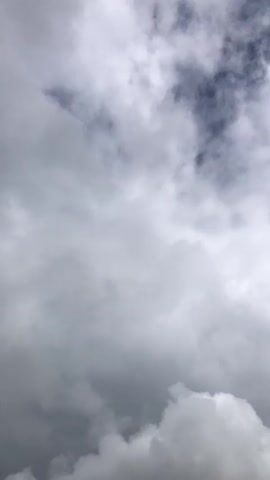 The video is a close-up, vertical shot of a cloudy sky. The clouds are a mix of white and gray, with patches of dark blue sky visible in between. The clouds appear to be moving, but the movement is subtle and not clearly defined. There are no people or objects visible in the video. The lighting suggests it is daytime.