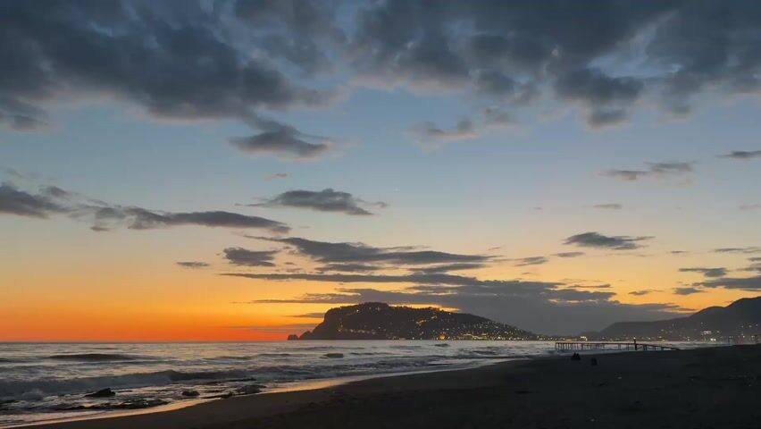 The video shows a scenic view of a beach at sunset. The sky transitions from a bright orange on the horizon to a blue hue higher up, with scattered dark clouds. The ocean waves gently roll onto the sandy beach. In the distance, a small, hilly island is visible, dotted with city lights. A few silhouetted figures can be seen on the beach. A pier-like structure extends slightly into the ocean on the right side of the frame.