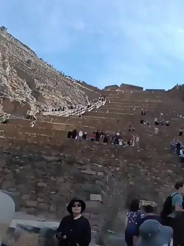 The video shows a crowded scene at an ancient stone terraced structure, possibly a historical site. Many people are walking on the steps of the terrace, while others are sitting or standing on different levels, observing the scenery. The sky is mostly clear, and a steep, rocky cliff rises behind the terraces. The people in the scene are dressed in casual clothing, and some are wearing hats or carrying backpacks. The overall atmosphere is that of a busy tourist attraction.