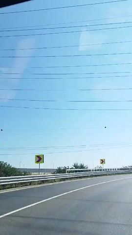 The video shows a perspective from inside a vehicle driving on a highway. The sky is clear and blue with some thin clouds. A red semi-trailer truck is visible driving in the same direction in the left lane. Several large metal electricity pylons and power lines are present along the side of the highway. Yellow road signs indicating curves are also visible. The road is paved and has white lane markings. The overall environment appears to be a sunny day on a highway.