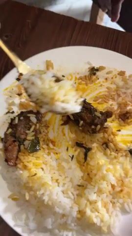 The video presents a close-up view of a white plate holding a meal, observed from an overhead perspective. The food comprises white rice, mixed with yellow-orange spiced rice, and several pieces of dark brown meat seasoned with green herbs. A gold-colored fork is actively engaged in scooping and mixing the food on the plate, indicating a person is in the process of eating.

The plate is placed on a wooden table featuring a visible grain and vertical plank design. In the background, a dark-skinned leg and foot, wearing brown sandals or flip-flops, are visible on a light-colored tiled floor. The activity central to the video is the consumption of the meal. The scene depicts an indoor setting, likely a dining area in Dushepe, Nigeria. No specific time of day, weather conditions, or emotions are discernible, and there is no visible text.