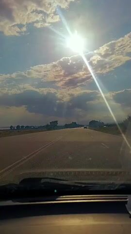 The video shows a first-person perspective from inside a vehicle traveling on a paved road. The sun is shining brightly in a partly cloudy sky, casting lens flares into the camera. There are a few other vehicles on the road ahead and to the side. The landscape appears to be rural with scattered trees and low vegetation visible in the distance. The interior of the car is partially visible in the foreground, including the dashboard and windshield.
