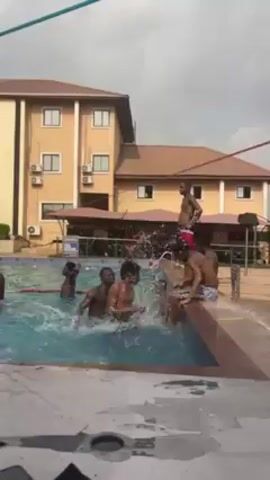 The video shows a group of young men in and around a swimming pool. Some are in the water, splashing and playing, while others are sitting or standing at the edge of the pool. A building is visible in the background. A sign with "Heineken" is visible behind the pool. The sky is cloudy.