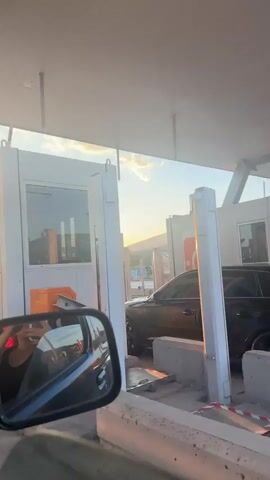 The video, shot from inside a car, shows the car passing through a toll booth at sunset. The booth is a white structure with orange accents. The sun is low in the sky, creating a golden glare. The car's side mirror is visible in the foreground.  The car stops briefly, likely to pay the toll, and then proceeds through. The sky is blue with scattered clouds. There is machinery behind the booth. A contrail is visible in the sky.