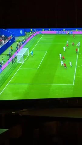 A video of a soccer game on a television screen. A goalkeeper in blue is in the penalty box. Several players in red and white uniforms are on the field, with the ball near the goal. Spectators are visible in the stands. The game appears to be in progress.