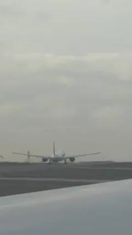 From inside an airplane, another plane can be seen in the distance taking off. The sky is cloudy and gray. The runway is visible in the foreground and background.