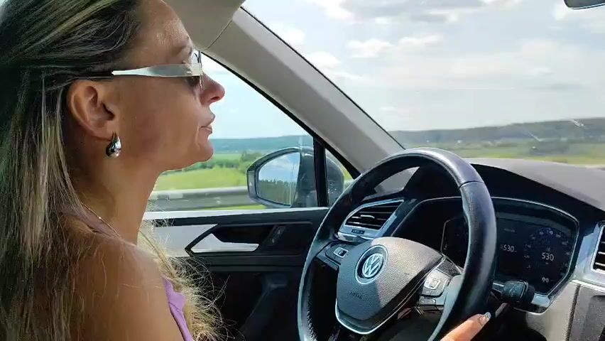 A woman with blonde hair and sunglasses is driving a Volkswagen. She is wearing a purple top and silver earrings. The car's interior is visible, including the steering wheel and dashboard. The car is on a road with trees and fields in the background. The sky is partly cloudy.