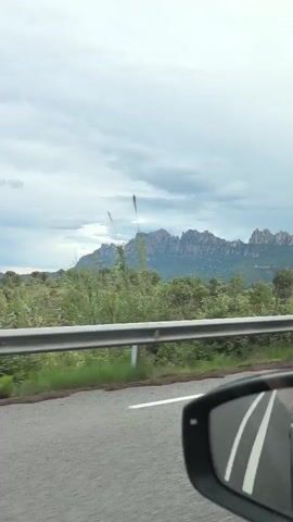A car is driving down a road with a mountain range in the background. The sky is overcast. There is a guardrail between the road and the scenery. The car's side mirror is visible. Upbeat music plays in the background.