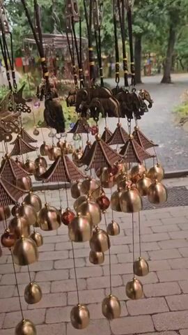 The video shows a close-up, eye-level view of several wind chimes hanging from a metal rack. The wind chimes are made of brass and have various decorative elements, including elephants, pagodas, and what appear to be horses. They are hanging outdoors, with trees and foliage visible in the background. The ground is paved with bricks. The camera is moving slightly, and the wind chimes are swaying, creating a gentle tinkling sound. The overall impression is one of a peaceful, outdoor market or garden setting.