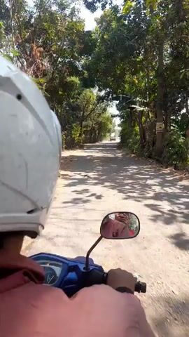 A first-person perspective video shows someone riding a blue and white scooter down a dirt road lined with trees and foliage. The rider is wearing a white helmet and a coral pink shirt. The rider’s left hand is visible on the scooter’s handlebar, and their right hand is also visible. The scooter’s rearview mirror reflects the rider’s face. Sunlight filters through the trees, casting shadows on the road. In the distance, another vehicle, possibly a white car, is visible on the road. The scene suggests a rural or tropical environment.