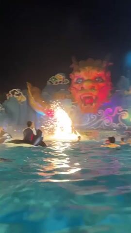 A nighttime view from a pool shows a performance with a large dragon-themed backdrop. In the foreground, a person is on the back of a dolphin or orca in the water, and several other people are in the pool, some with flotation devices. A fountain of water is spraying upwards, with fireworks creating a bright explosion of light and sparks above the water, partially obscured by mist and smoke. The dragon backdrop is illuminated with colorful lights, and the overall scene appears to be an entertainment show.