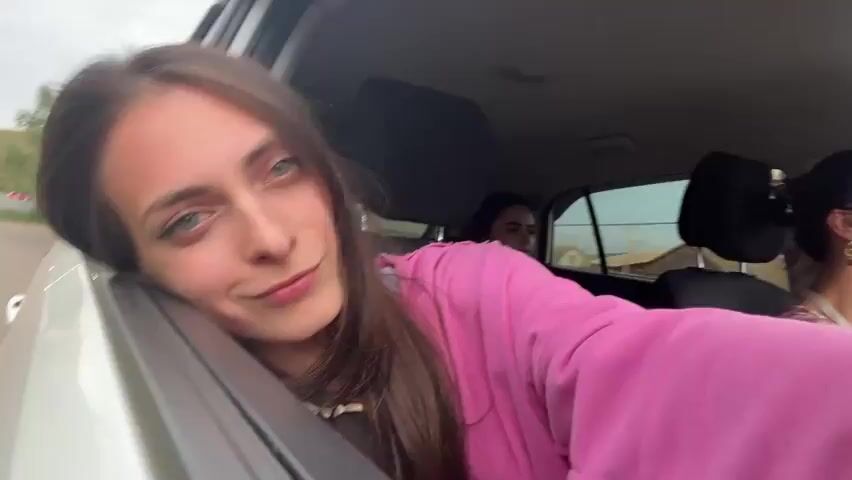 The video shows a young woman with long brown hair leaning out of a car window and looking towards the camera. She is wearing a bright pink sweatshirt. Her head is resting on the car door, and she has a playful expression on her face. In the background, another woman is visible in the back seat of the car, and the road and trees are passing by outside the window. The woman in the foreground smiles and waves her hand out of the window, appearing to be enjoying the ride. The video is shot from a first-person perspective, as if the viewer is the one holding the camera.
