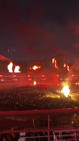The video captures a concert event at night, held in a large stadium filled with a massive crowd of people.  The stage is set against the backdrop of a giant video screen displaying images of a face and fiery visual effects. There are pyrotechnic displays going off around the stage, sending flames shooting into the air. The scene is lit up with bright stage lights, with a predominant red hue. The camera is positioned from a higher vantage point, possibly a balcony or elevated seating area, overlooking the stage and the audience.
