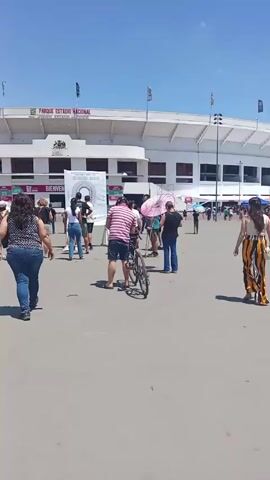 Here is a description of the video:
The video shows a flat, open plaza in front of a stadium on a sunny day. The stadium, labeled "Parque Estadio Nacional," is white with a horizontal band of red and black. Flags are visible above the stadium. Several people are walking around the plaza. A man stands with a bicycle near the center of the frame. To the left, a woman in jeans and a printed shirt walks away from the camera. Further along, a group of people gathers around a white poster with a black-and-white image. One man among the group opens a pink and white parasol. A woman wearing a long, striped skirt and a black top walks across the plaza. Other individuals are seen in the background, some near what appear to be tents.