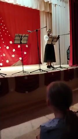 A person stands on a stage in front of a music stand. They are dressed in a white blouse and a black skirt. The stage has a red and white backdrop and red curtains. There are microphones and more music stands. A person is visible in the foreground of the shot in the audience.
