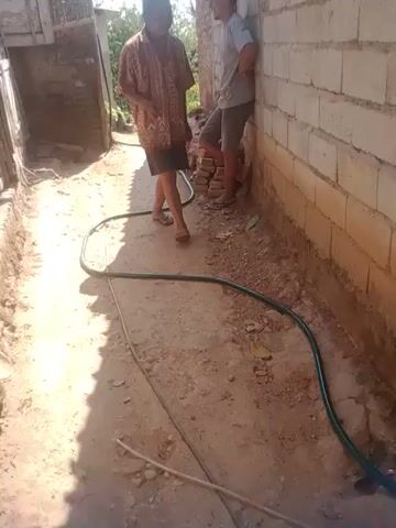 The video shows a person standing next to a brick wall. The person is wearing a patterned shirt and dark shorts. They are holding a green hose and appear to be watering the ground. The ground is dirt and gravel. There are some plants visible in the background. The video is filmed outdoors in daylight.