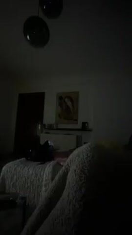 The video depicts a dim interior space, likely a living room or bedroom, with a sofa in the foreground covered with a textured blanket and cushions. In the distance, a shelf or sideboard holds decorative objects beneath a framed picture on the wall. To the left, a dark doorway is visible. As the camera zooms slightly, a white and dark-furred cat comes into clearer view, resting on the blanket-covered sofa. The overall lighting is low, making details difficult to discern.