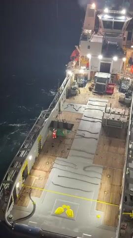This media file captures a nighttime scene on the deck of a large vessel, likely a ship or a platform operating at sea. The primary subjects are two individuals engaged in an activity involving a large, green skip or dumpster. One person, wearing a bright red and white protective suit, stands near the skip, appearing to direct or assist in its loading. Another individual is partially visible inside or directly above the skip, also seemingly involved in the task.

The setting is the open deck of the vessel, characterized by weathered wooden planks and a gray, non-slip surface. Several thick ropes are coiled and laid out on the deck, suggesting preparedness for maritime operations. Various pieces of equipment, including what appears to be a winch or a generator, are visible further up the deck. The vessel itself has a white superstructure with numerous lights illuminating the work area, indicating nighttime operation.

The activity taking place is the handling and potential loading or unloading of the skip. The presence of the skip and the individuals' attire suggest a work environment, possibly related to waste management or material transfer. The dark water surrounding the vessel and the choppy waves indicate a marine environment.

Notable details include the time of day, clearly nighttime due to the artificial lighting and the dark surroundings. There are no clear weather indicators, though the presence of waves suggests a body of water. Location cues are limited to it being a marine vessel. Emotions are difficult to ascertain from the image alone, but the focused activity suggests a professional and operational mood.

No visible text is clearly legible in the provided frames. The overall impression is one of an industrial operation taking place at sea under the cover of darkness.