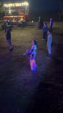 The video shows a nighttime outdoor scene, possibly a festival or concert, with a stage in the background lit by bright lights. In the foreground, a person with dreadlocks in their hair, wearing a colorful patterned jacket and brightly colored pants, is dancing with their arms outstretched. Several other people are visible in the background, standing and observing. The ground is covered in dirt and grass. The lighting suggests artificial lights from the stage and possibly other sources, creating shadows and illuminating parts of the scene with purple and pink hues.