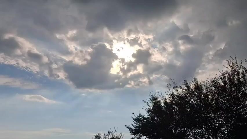 The video shows a cloudy sky with the sun peeking through. The clouds are dark and dramatic, with rays of light shining through them. There are trees visible in the foreground, silhouetted against the sky. The video appears to have been filmed outdoors, possibly on a breezy day.