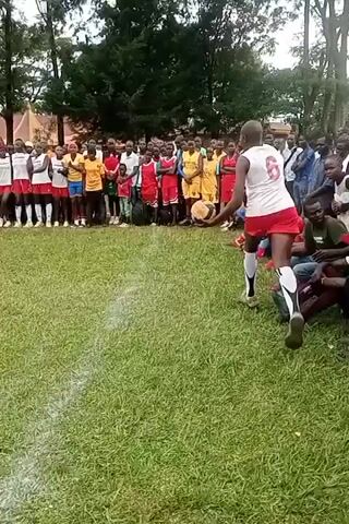 The video shows an outdoor volleyball game on a grass field. A crowd of people lines the edges of the field, watching the game. The volleyball net is stretched across a bare patch of ground. Players wearing white jerseys with red shorts and numbers on their backs are playing against a team that is not entirely visible. A player serves the volleyball over the net.