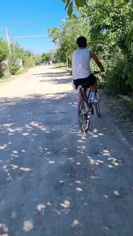 A person is riding a bicycle down a dirt road lined with trees. The person is wearing a white tank top and dark shorts. The sky is blue and there are some clouds. The road is bumpy and there are shadows from the trees on the road. The camera is positioned behind the person, following them as they ride.
