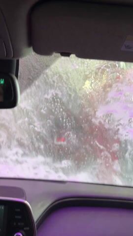 The video shows the inside of a car going through a car wash. Pink and green soap and water cover the windshield and sunroof. The car's interior is visible.